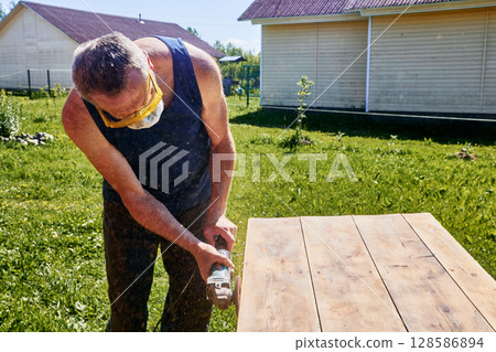 Holding angle grinder with both hands, craftsman wearing respirator and safety glasses sanding tabletop outdoors in rustic countryside setting 128586894