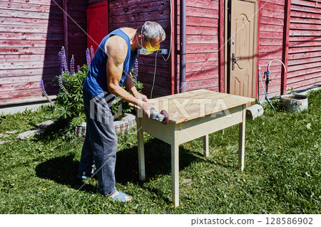Sanding wooden edge with handheld grinder, carpenter wearing respirator and safety glasses working outdoors on tabletop in rural surroundings 128586902