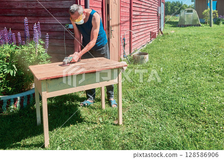 Grinding wooden tabletop with angle grinder, handyman wearing respirator and safety glasses working outdoors in rural environment. 128586906