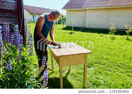 Sanding wooden tabletop with angle grinder outdoors, hobbyist working in yard near wooden houses on sunny summer day in countryside. 128586908