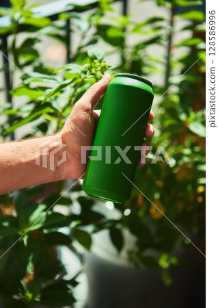 Holding blank green beverage can with lush plants backdrop for advertisement mockup 128586996