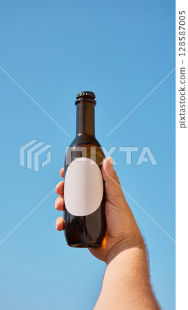 Hand grasping bottle with blank white label against a clear blue sky for advertising mockup 128587005