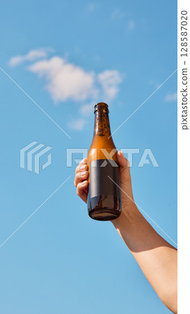 Hand holding beer bottle with blank label in front of clear blue sky with white clouds Hand holding beer bottle with blank label in front of clear blue sky with white clouds 128587020
