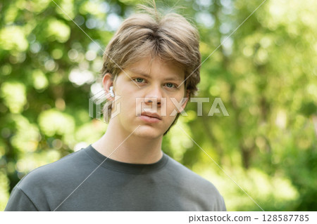 Portrait of strict young man standing outdoors with earbuds in public park setting, looking at camera. Summer season and green foliage as background creates serene atmosphere. Close up. 128587785