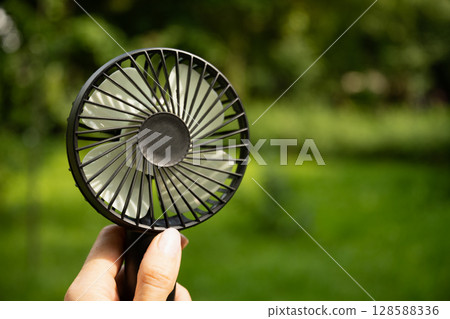 Electric fan in woman hand suffering from heat at park. Overheated suffering from heat attack uses an electric handy fan to cool himself down in hot summer day. Beat heat. Global warming 128588336