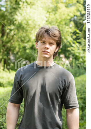 Portrait of young blond man standing outdoors with earbuds in public park setting, looking at camera. Summer season and green foliage as background. 128588339