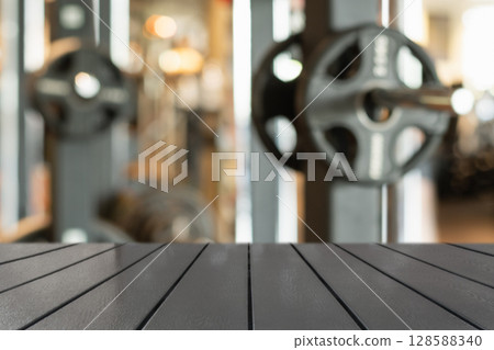 Wooden table surface in foreground with blurred gym background featuring weightlifting equipment. Fitness and healthy lifestyle themes. Space for design. Wooden table surface in foreground with blurred gym background featuring weightlifting equipment. Fitness and healthy lifestyle themes. Space for design. 128588340