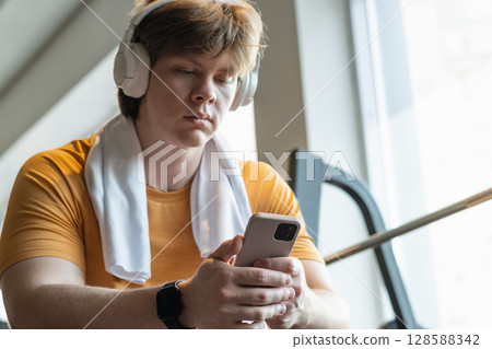 Sporty young man relax and sitting with mobile phone, listen music headphones and texting at gym. Bodybuilder during training . Fitness lifestyle concept. Take a break, looking at camera. Sporty young man relax and sitting with mobile phone, listen music headphones and texting at gym. Bodybuilder during training . Fitness lifestyle concept. Take a break, looking at camera. 128588342