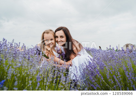 A young woman in a long white dress gently hugs her little daughter in the middle of a lavender field 128589066