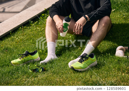 Man resting on grass with green running shoes and water bottle after exercise Man resting on grass with green running shoes and water bottle after exercise 128589601