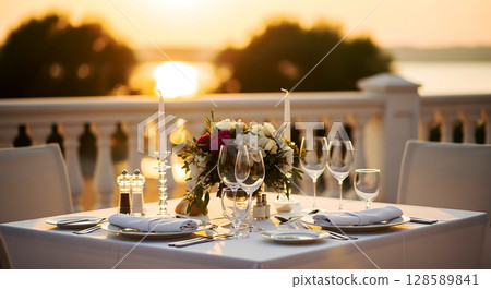 Elegant dining setup with candles, flowers, wine glasses, and cutlery on a white tablecloth against a sunset view over water. Soft light highlights a romantic atmosphere Elegant dining setup with candles, flowers, wine glasses, and cutlery on a white tablecloth against a sunset view over water. Soft light highlights a romantic atmosphere 128589841