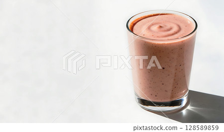 A pink, creamy smoothie with visible berry specks swirled evenly in a transparent glass. It casts a soft shadow on a light surface, suggesting natural daylight illuminating it 128589859