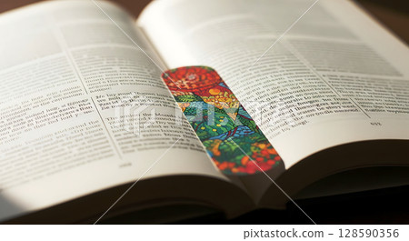 Open book with colorful patterned bookmark between pages, sunlight highlights printed text and shadows cast on the pages, creating a warm, inviting atmosphere for reading 128590356
