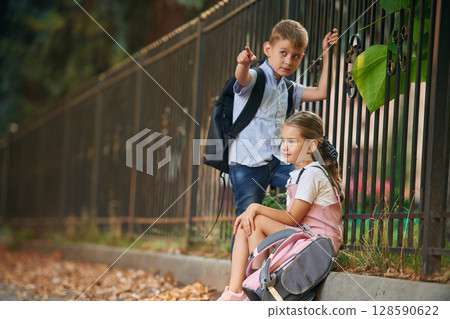 By the fence, relaxing. Young school children of boy and girl are together outdoors By the fence, relaxing. Young school children of boy and girl are together outdoors 128590622