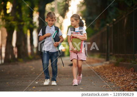 Front view, walking together. Young school children of boy and girl are outdoors Front view, walking together. Young school children of boy and girl are outdoors 128590629