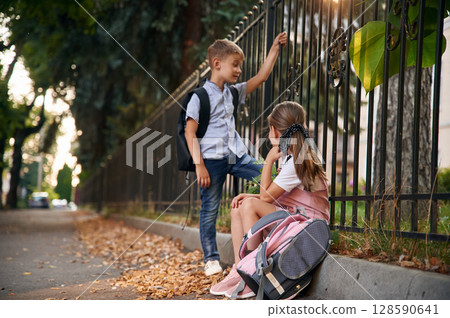 By the fence, relaxing. Young school children of boy and girl are together outdoors By the fence, relaxing. Young school children of boy and girl are together outdoors 128590641