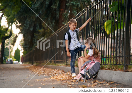 By the fence, relaxing. Young school children of boy and girl are together outdoors By the fence, relaxing. Young school children of boy and girl are together outdoors 128590642