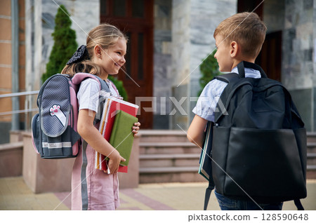 Holding notebook and books. Young school children of boy and girl are together outdoors Holding notebook and books. Young school children of boy and girl are together outdoors 128590648