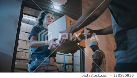 Outside of Logistics Retail Warehouse at Night: Female Worker Giving Cardboard Box to Colleague 128590936
