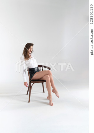Middle-aged woman posing on a chair in a bright studio, dressed in a white shirt and black shorts. Concept: Reflecting the timeless appeal of classic wardrobe staples and their modern interpretations. Middle-aged woman posing on a chair in a bright studio, dressed in a white shirt and black shorts. Concept: Reflecting the timeless appeal of classic wardrobe staples and their modern interpretations. 128591159