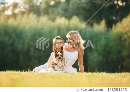 Sitting on the ground. Mother with her daughter and cute dog are on the field outdoors 128591255