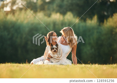 Sitting on the ground. Mother with her daughter and cute dog are on the field outdoors 128591256