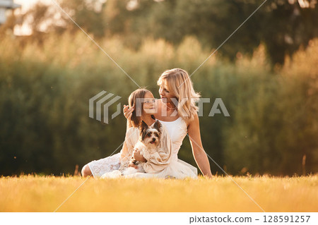 Sitting on the ground. Mother with her daughter and cute dog are on the field outdoors 128591257
