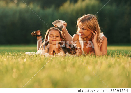 Lying down on the grass. Mother with her daughter and cute dog are on the field outdoors Lying down on the grass. Mother with her daughter and cute dog are on the field outdoors 128591294