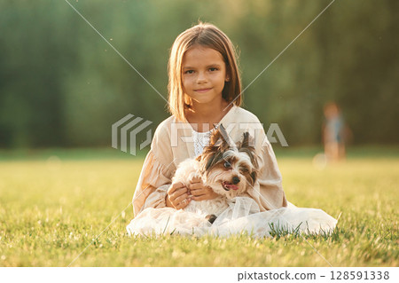 Front view, sitting together. Cute little girl is on the field with dog 128591338