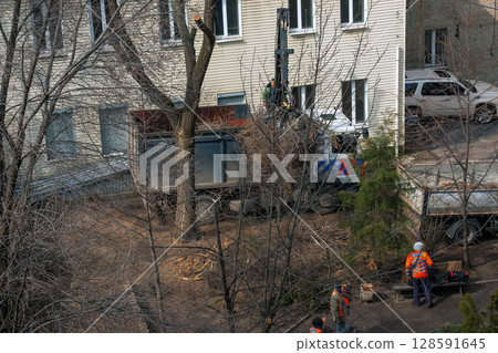 Dnipro, Ukraine - March 20, 2024: City utility workers use bulldozer to remove tree branches after pruning in the courtyard of an apartment building. 128591645