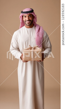 Smiling middle-eastern adult male holding wrapped gift box on beige background. 128592183
