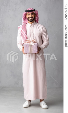 Smiling middle-eastern young male in traditional attire holding gift box. 128592189