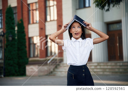 Playing with book that is over the head. School girl in uniform is outdoors near the building Playing with book that is over the head. School girl in uniform is outdoors near the building 128592561