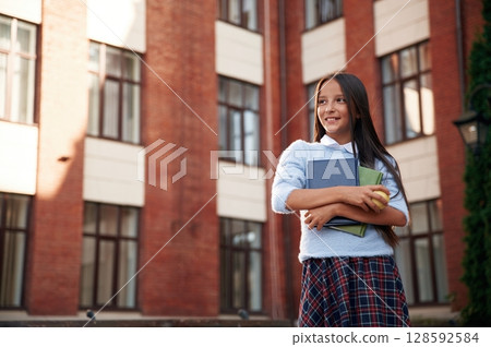Positive emotions, standing and holding books. School girl in uniform is outdoors near the building Positive emotions, standing and holding books. School girl in uniform is outdoors near the building 128592584