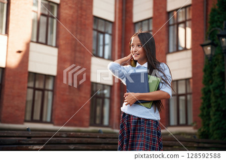 Positive emotions, standing and holding books. School girl in uniform is outdoors near the building 128592588