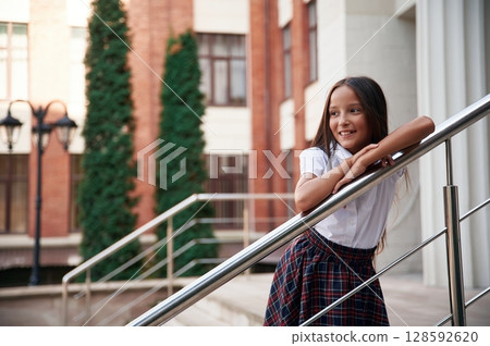 Leaning on the railing. School girl in uniform is outdoors near the building 128592620