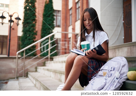 With backpack, sitting on the stairs. School girl in uniform is outdoors near the building With backpack, sitting on the stairs. School girl in uniform is outdoors near the building 128592632