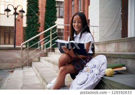 Having free time, sitting on the stairs. School girl in uniform is outdoors near the building 128592633