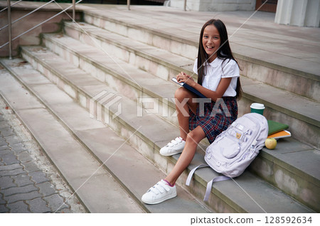 Having free time, sitting on the stairs. School girl in uniform is outdoors near the building Having free time, sitting on the stairs. School girl in uniform is outdoors near the building 128592634