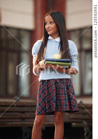 With notepad in hands. School girl in uniform is outdoors near the building 128592691