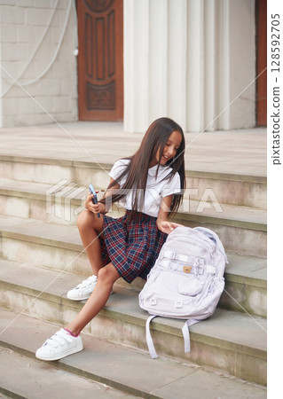 Having free time, sitting on the stairs. School girl in uniform is outdoors near the building Having free time, sitting on the stairs. School girl in uniform is outdoors near the building 128592705