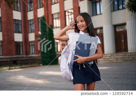 Front view. School girl in uniform is outdoors near the building Front view. School girl in uniform is outdoors near the building 128592714