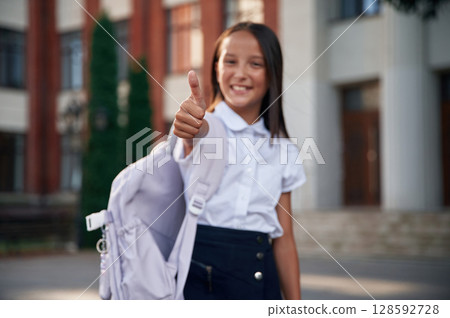 Showing thumb up. School girl in uniform is outdoors near the building Showing thumb up. School girl in uniform is outdoors near the building 128592728