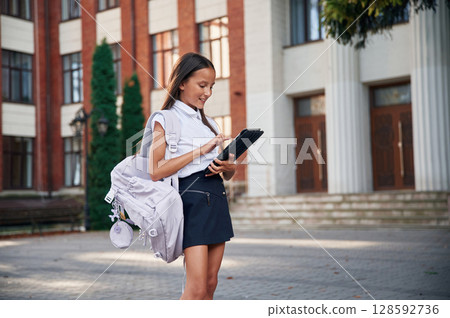 In classic uniform. School girl is outdoors near the building 128592736