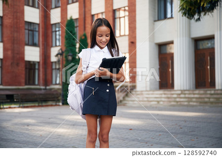 Wireless device in hands, standing. School girl in uniform is outdoors near the building 128592740