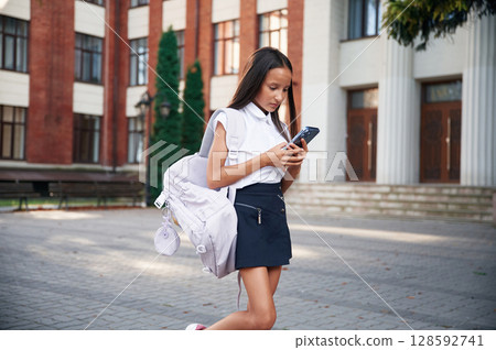 Wireless device in hands, standing. School girl in uniform is outdoors near the building 128592741