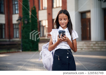 Phone in hands. School girl in uniform is outdoors near the building Phone in hands. School girl in uniform is outdoors near the building 128592749