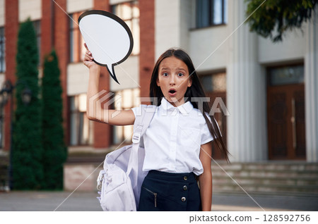 Sticker with thoughts, copy space for text. School girl in uniform is outdoors near the building 128592756
