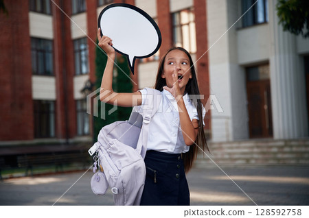Sticker with thoughts, copy space for text. School girl in uniform is outdoors near the building Sticker with thoughts, copy space for text. School girl in uniform is outdoors near the building 128592758