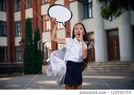 Sticker with thoughts, copy space for text. School girl in uniform is outdoors near the building Sticker with thoughts, copy space for text. School girl in uniform is outdoors near the building 128592759